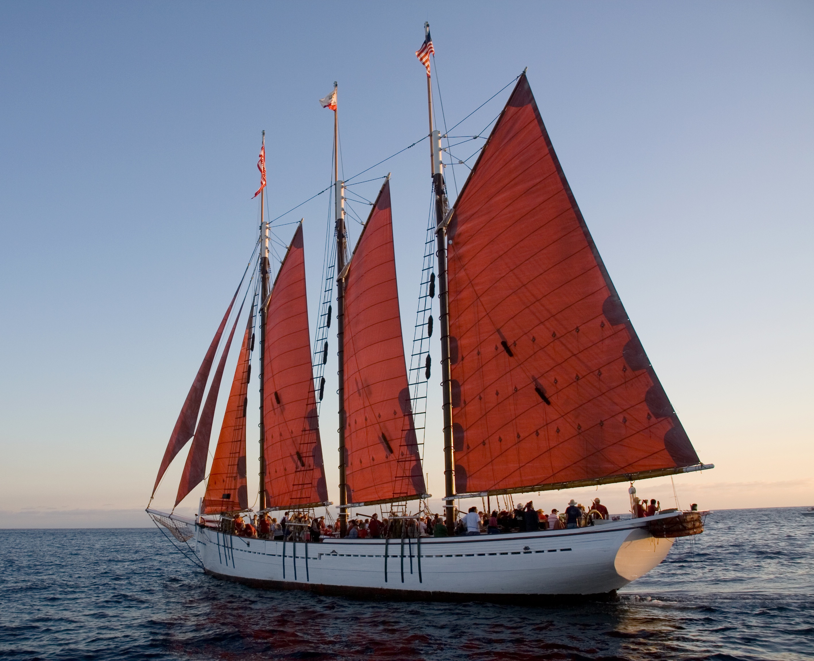 Wooden three-masted schooner under red sails at sunset